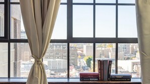 A window with a view of a cityscape, a curtain tied at the top, and a stack of books on the windowsill. - Sosuite 2BR Penthouse w W D Gym On-site Bar (Philadelphia)