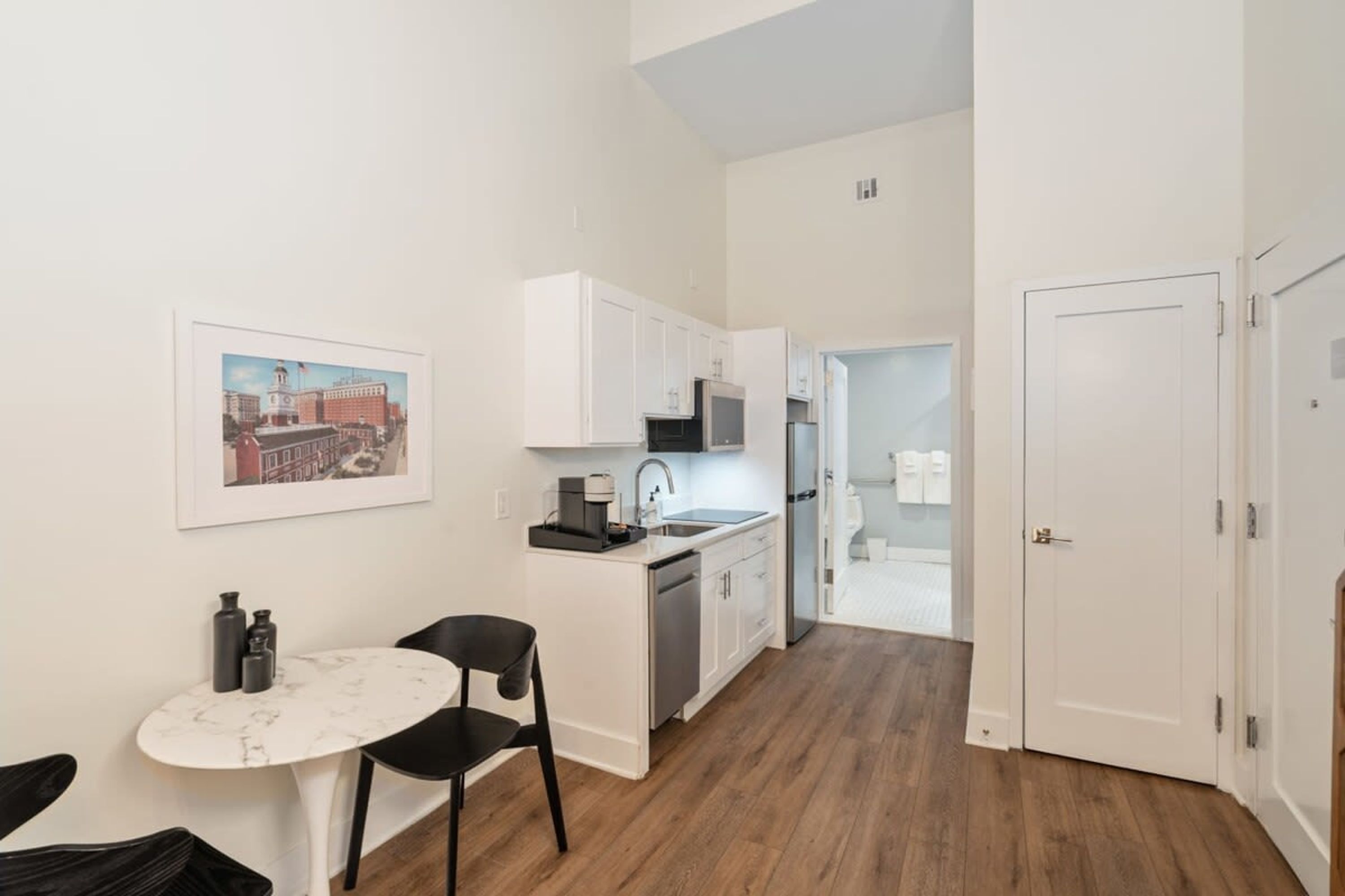 A compact kitchen with white cabinets, a small dining area with a marble table, and a door leading to a bathroom.
