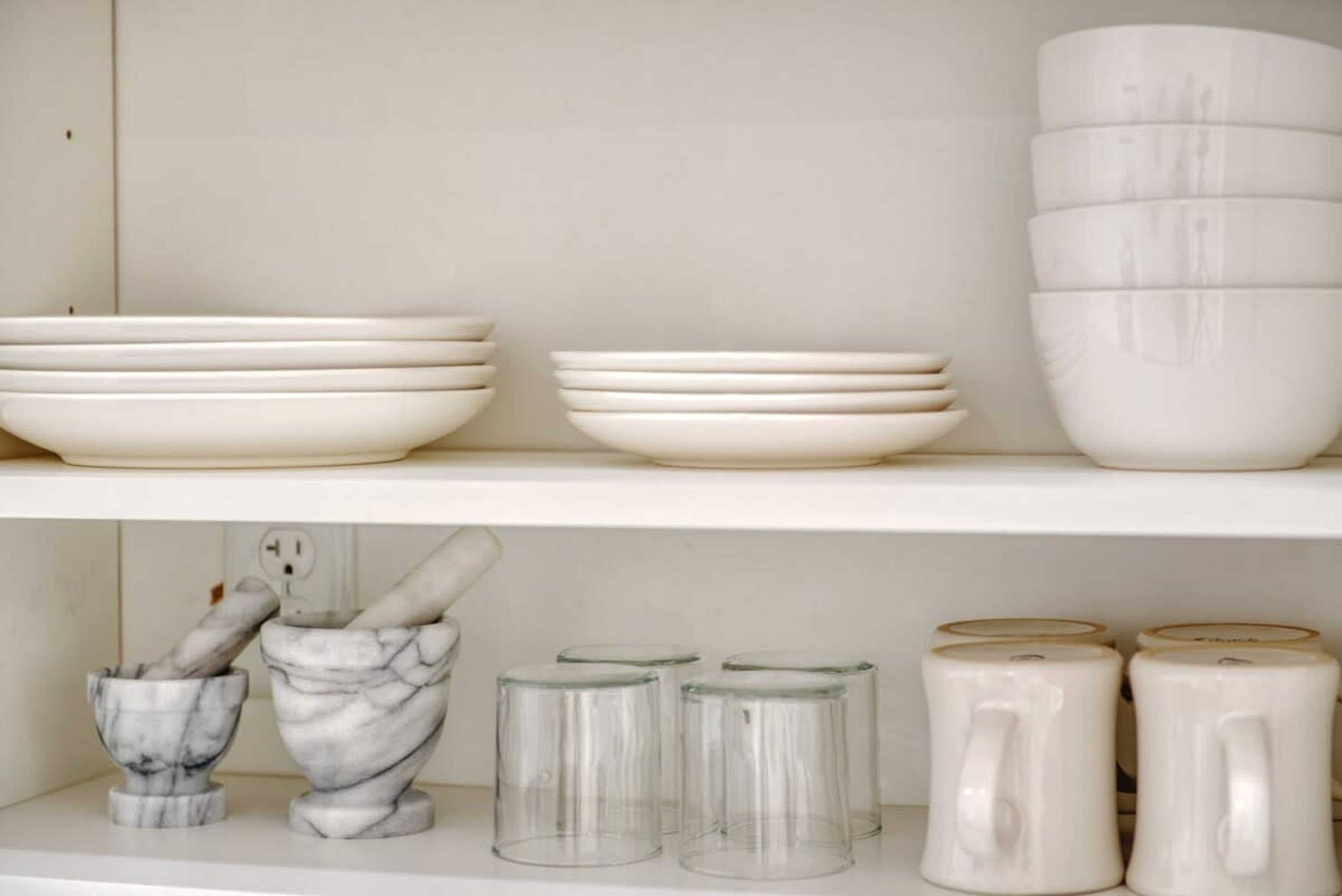 A shelf with white bowls, a marble mortar and pestle, and clear glass mugs.