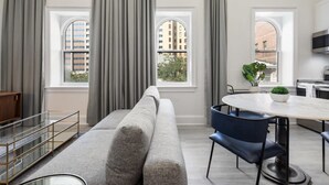 Een moderne woonkamer met een grijze bank, een eettafel met stoelen en grote ramen met gordijnen.