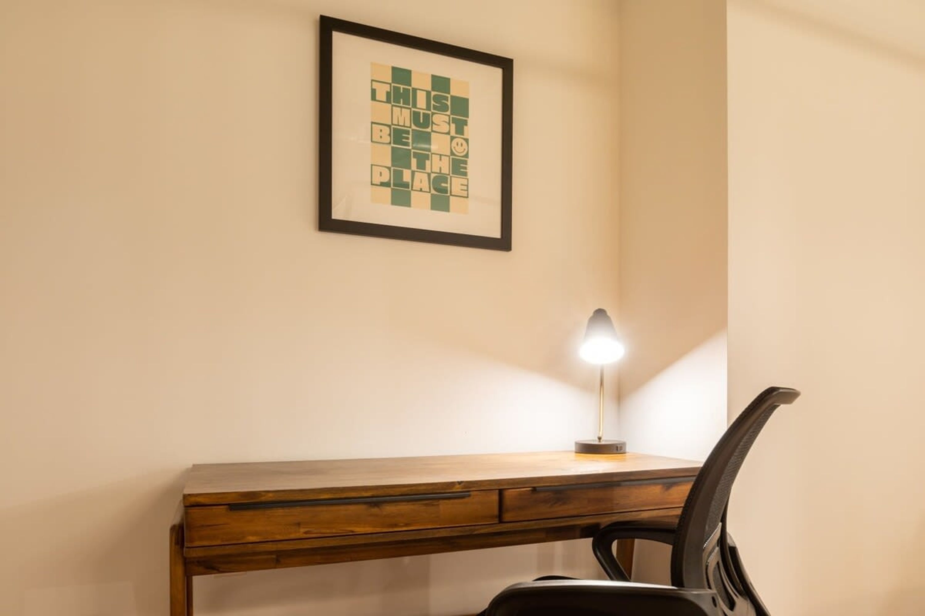 Un escritorio de madera con una lámpara, una obra de arte enmarcada y una silla negra.