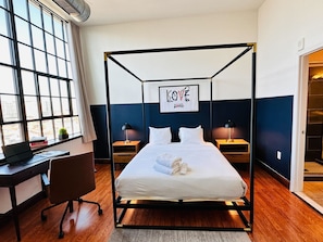 A modern hotel room with a four-poster bed, a desk with a laptop, a chair, and a large window offering a city view.