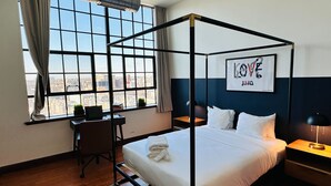 A four-poster bed with a city view, a desk with a chair, and a bedside table with a lamp.