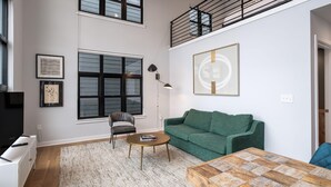 Een ruime woonkamer met een groene bank, een houten salontafel en een groot raam met jaloezieën.