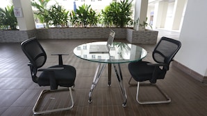 A round glass table with two black office chairs and a laptop on it, set on a tiled floor in a spacious indoor area with potted plants and a white building in the background. - Collection O 93750 Luxury Rooms @ Urbantown Serpong (South Tangerang)