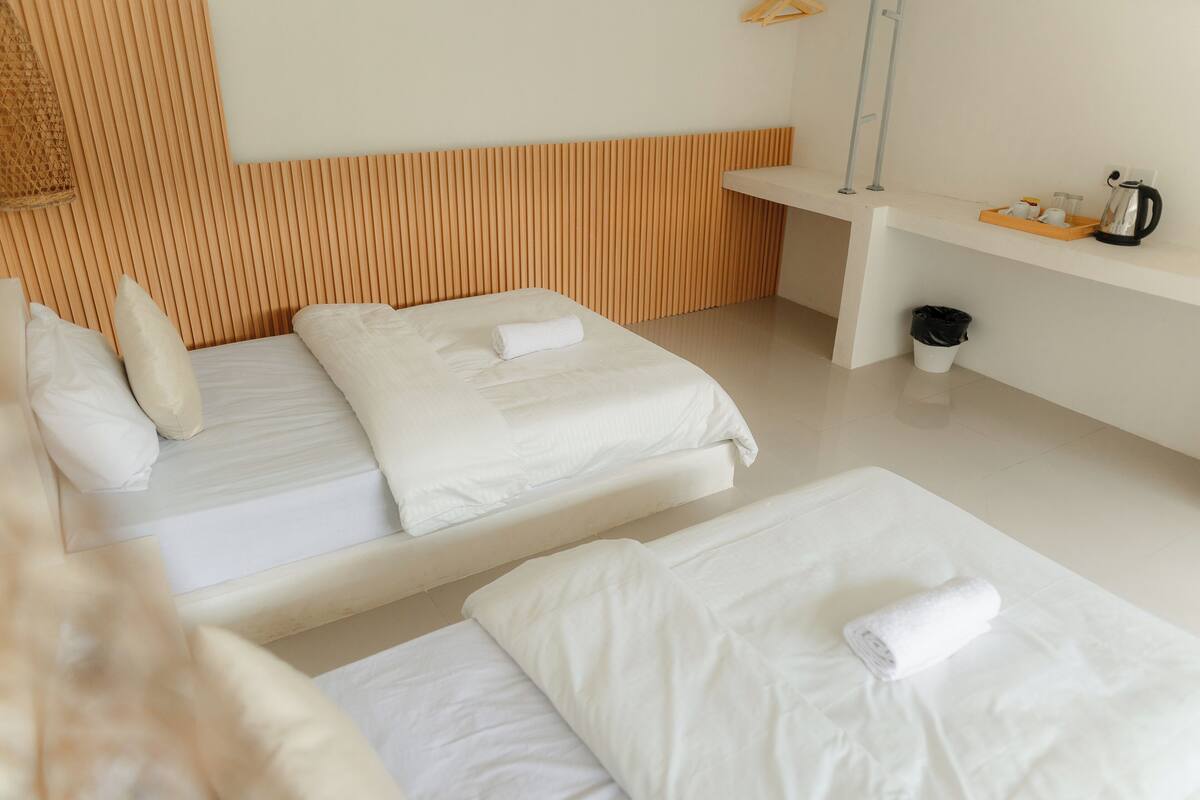 A hotel room with two beds, a wooden headboard, a small shelf with a kettle and tea set, and a trash bin.