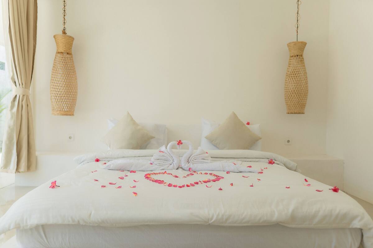 A neatly made bed with a heart-shaped flower arrangement and swan-shaped towels, two woven wall hangings, and a light-colored room with a curtain.