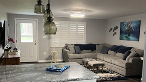 A modern living room with a sectional sofa, a marble kitchen island, and a large window with shutters.