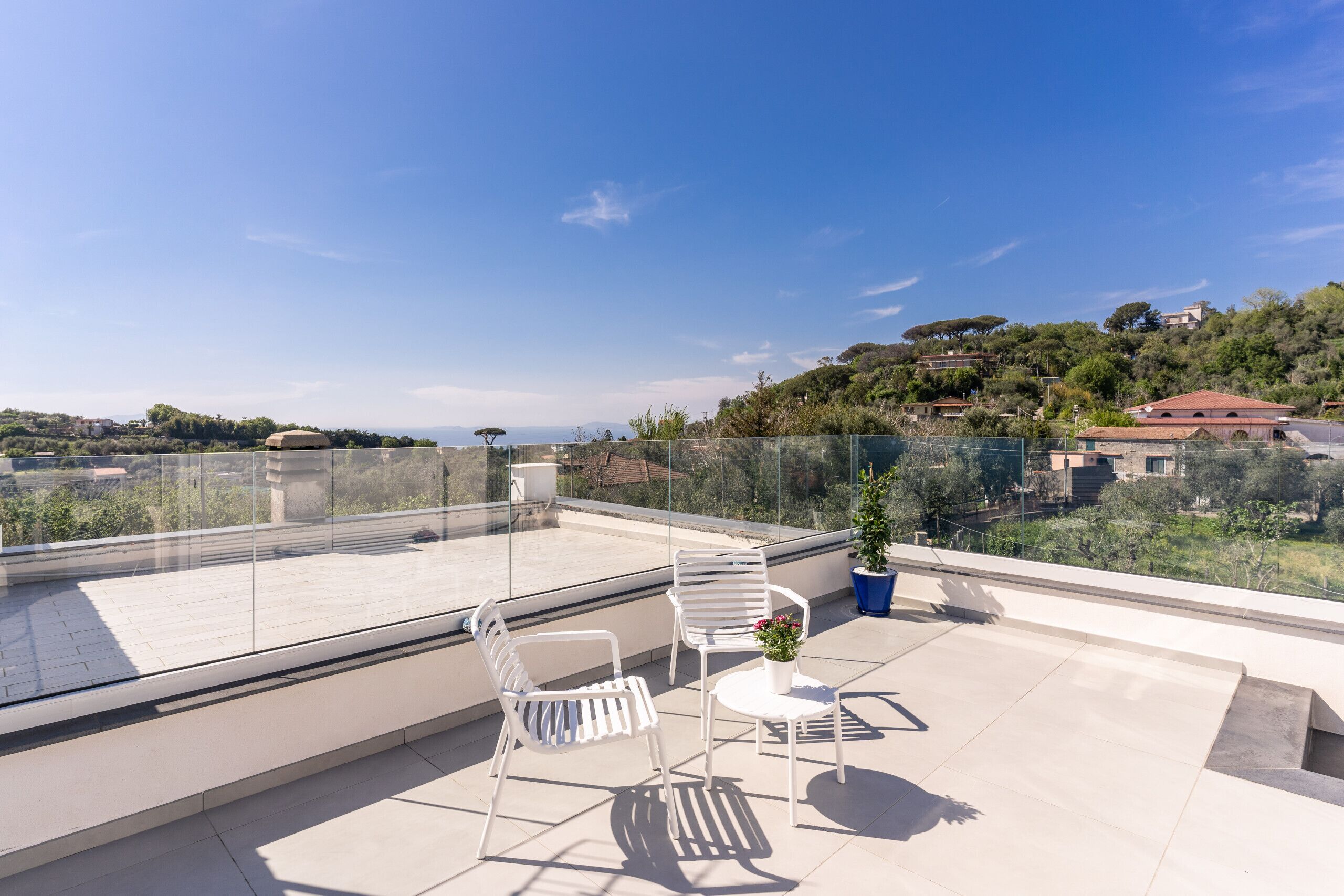 A rooftop terrace with two white chairs and a small table, a potted plant, and a clear view of a hilly landscape.