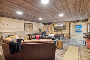 A log cabin interior with leather sofas, a stone fireplace, and a mounted television. - Cozy Lodge w/Theater|Firepit|Arcade|Hot Tub (Gatlinburg)