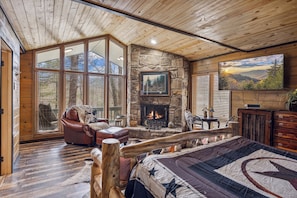 A cozy cabin interior with a stone fireplace, leather furniture, and a large window offering a view of the outdoors. - Cozy Lodge w/Theater|Firepit|Arcade|Hot Tub (Gatlinburg)