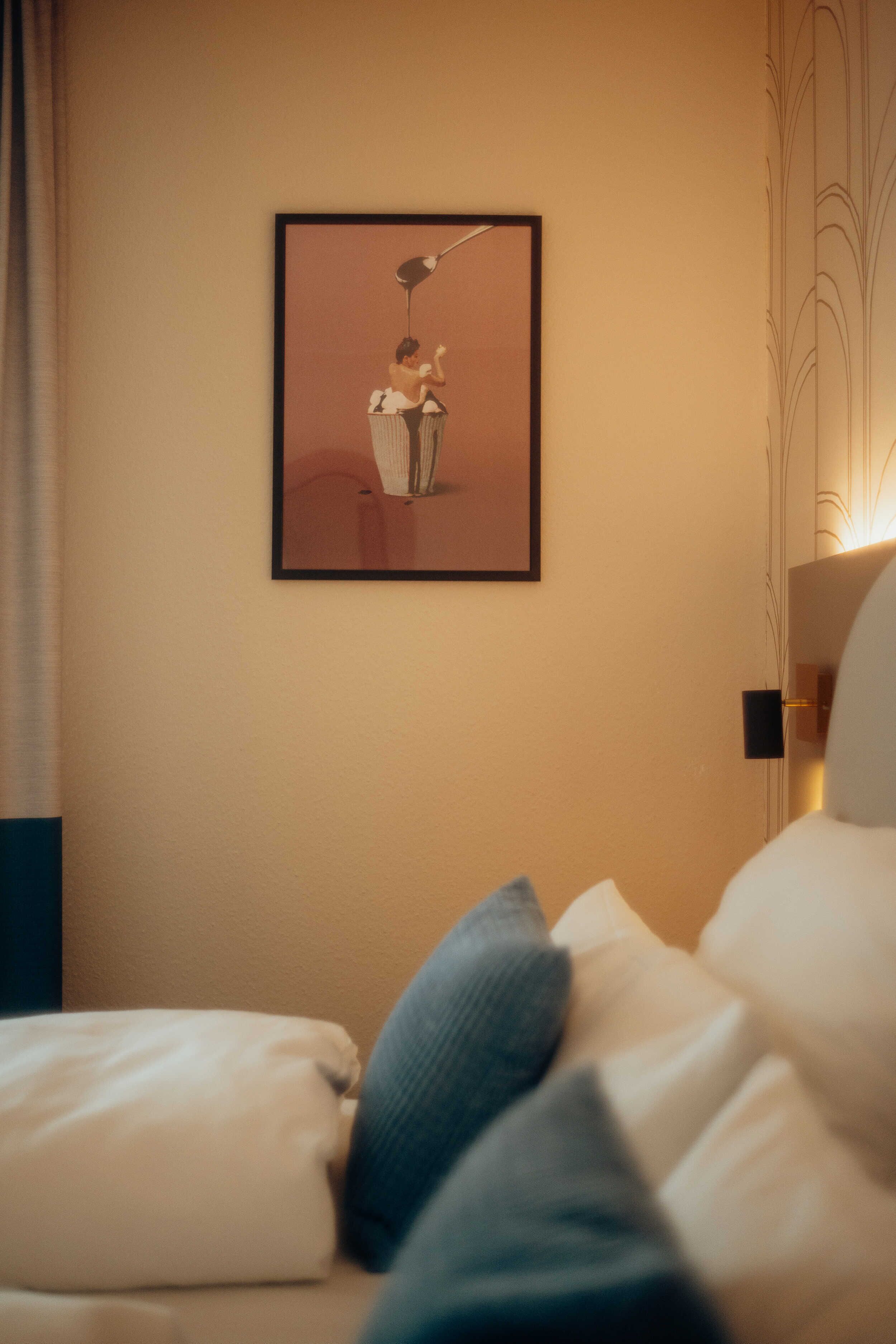 Ein Hotelzimmer mit einem gerahmten Kunstwerk an der Wand, einem Bett mit weißer Bettwäsche und einem blauen Kopfkissen sowie einem Nachttisch mit einer Lampe.