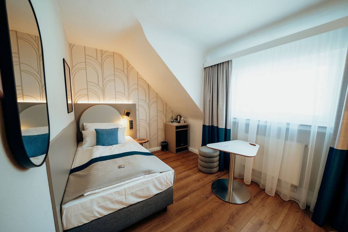 Ett modernt hotellrum med en stor säng, ett litet bord, en spegel och ett fönster med gardiner.