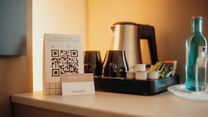 A QR code reminder sign on a hotel reception desk with a coffee maker, tea set, and water bottle. - guestified. LIYANA Hotel (Ratingen)