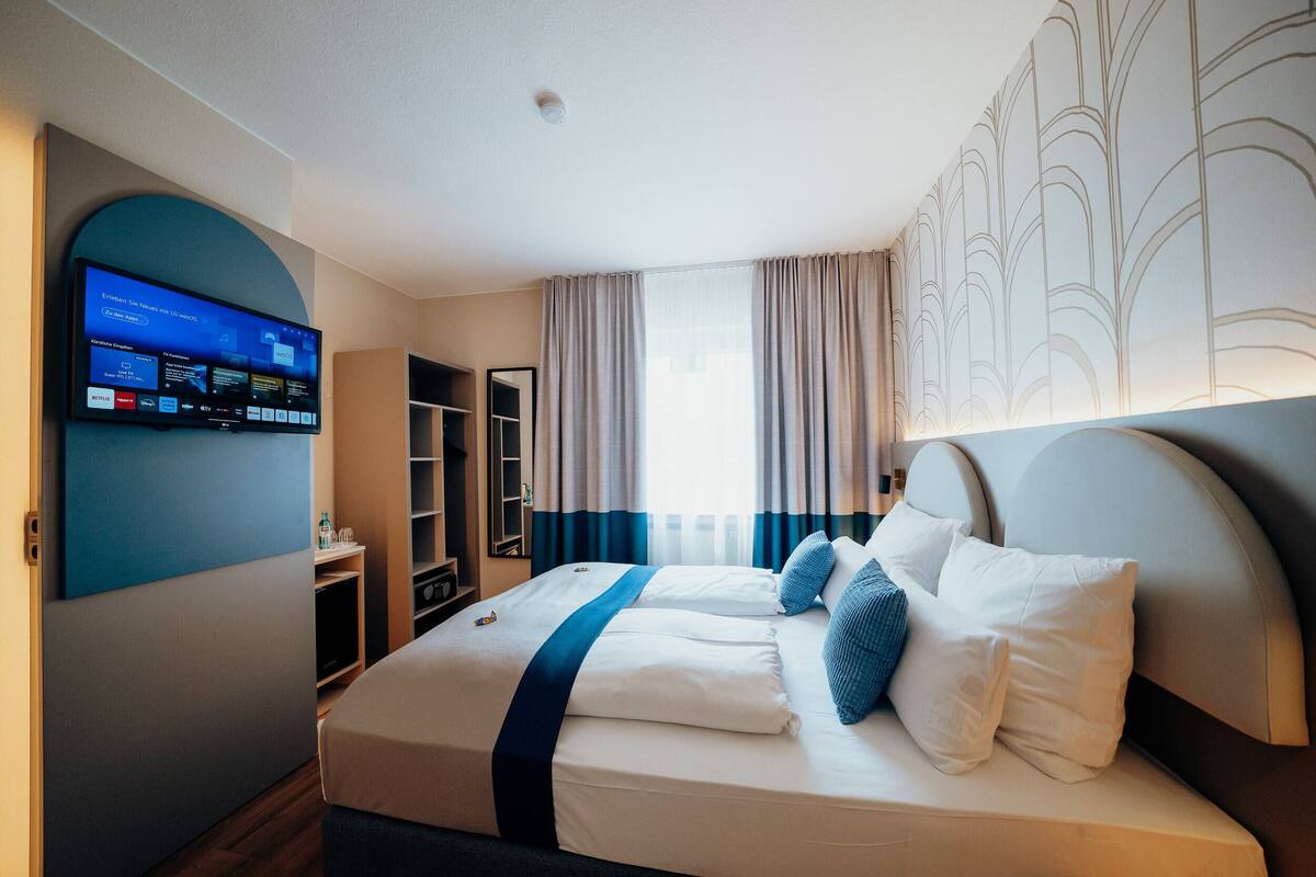 Ett hotellrum med en stor säng, en tv som är monterad på väggen, en garderob och ett fönster med gardiner.