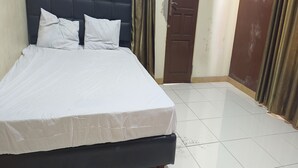 Un lit bien fait, avec du linge de lit blanc et une tête de lit sombre, dans une pièce dont le sol est recouvert de carreaux et la porte est marron.