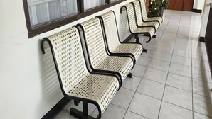 Une rangée de bancs en métal blanc, dotés d’une structure noire et d’un dossier perforé, est placée contre un mur, juste en dessous d’une fenêtre.