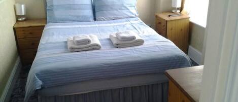 A neatly made bed with two folded towels, a wooden nightstand with a lamp, and a window with curtains.