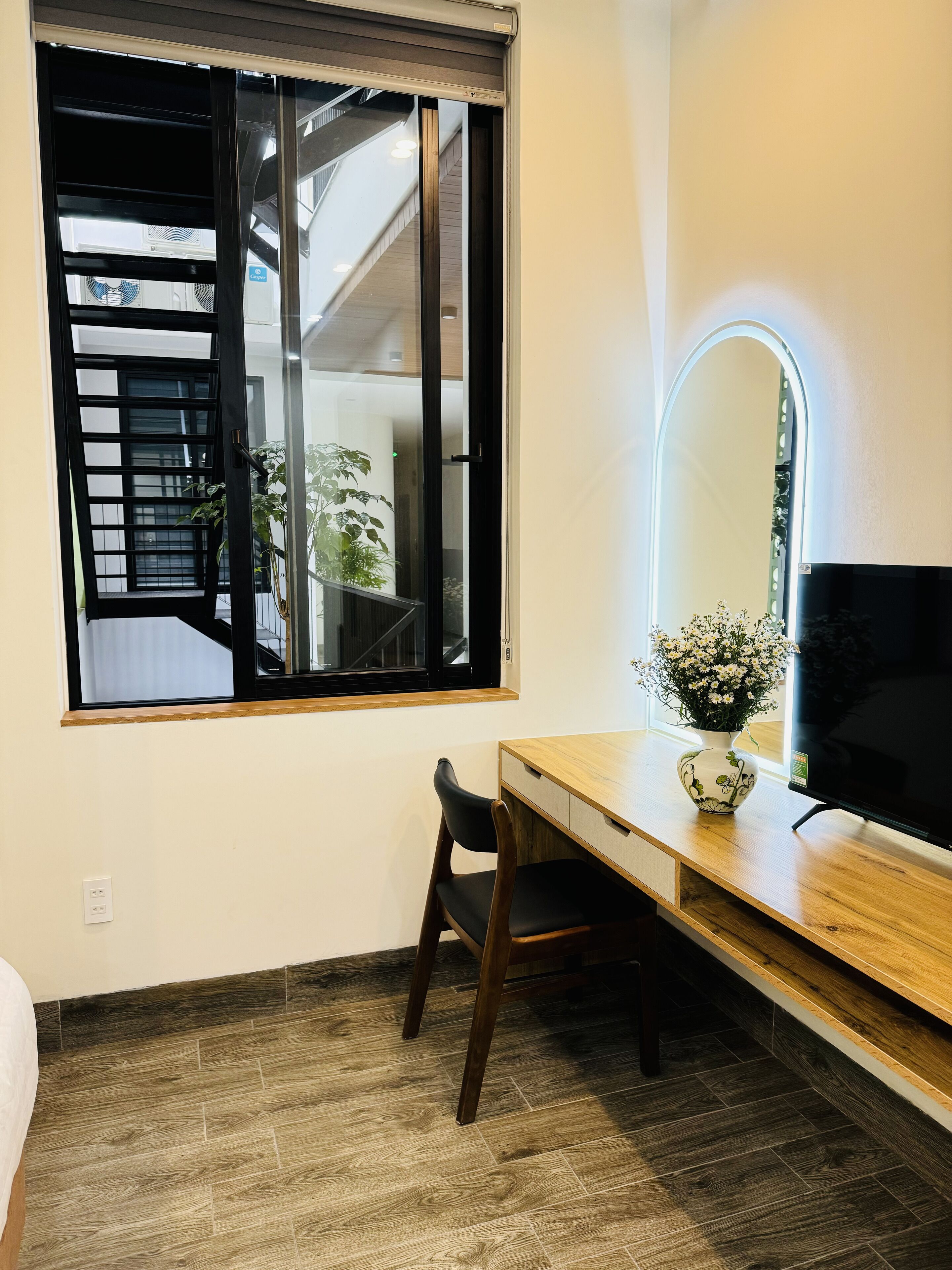 A room with a wooden desk, a chair, a TV, a vase with flowers, and a window with a view of a staircase and plants.