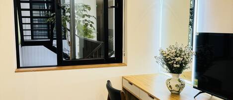 A room with a wooden desk, a chair, a TV, a vase with flowers, and a window with a view of a staircase and plants.