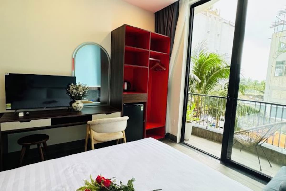 A hotel room with a bed, a TV, a desk with a computer, a red wardrobe, and a balcony with a view of palm trees.