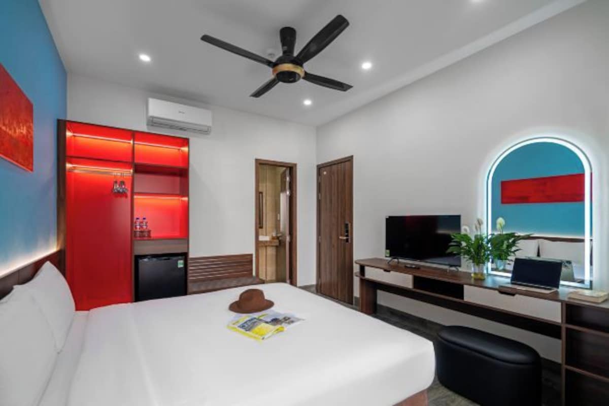 A hotel room with a bed, a desk with a TV, a ceiling fan, and a red-lit wardrobe.