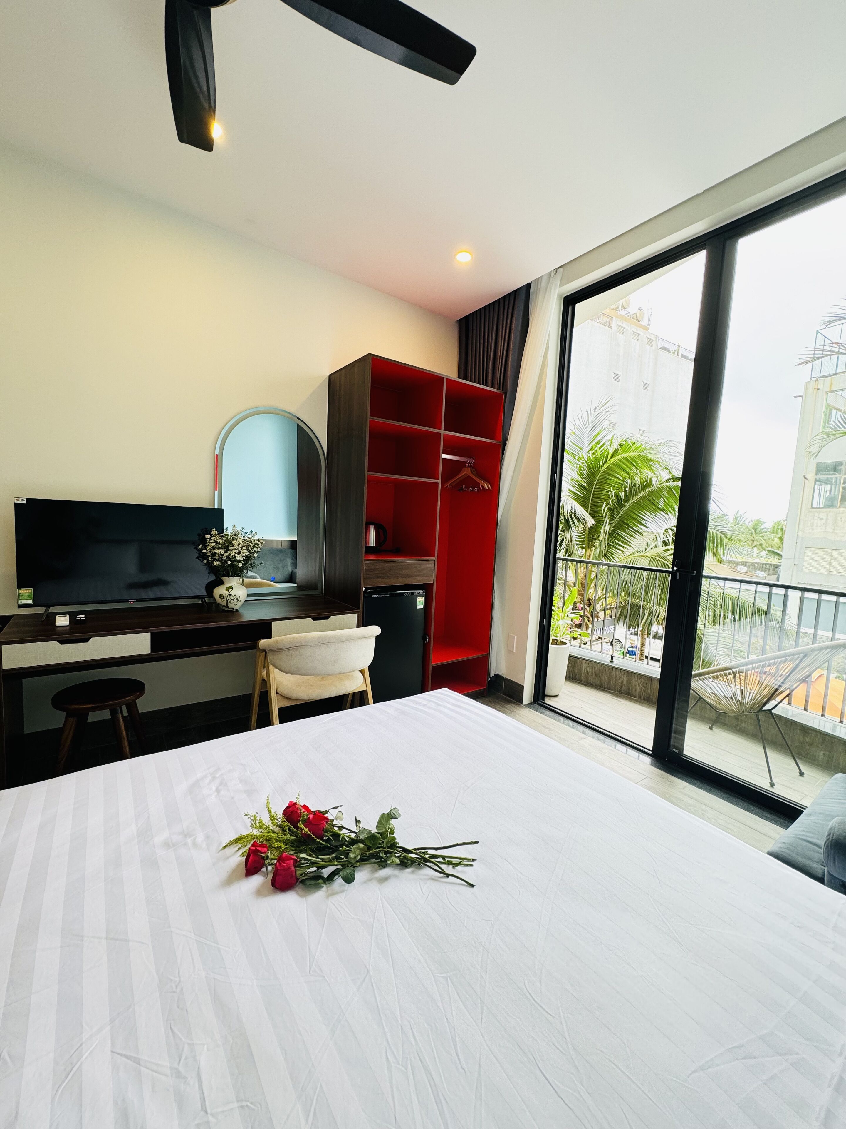 A hotel room with a bed, a desk with a TV, a red wardrobe, and a balcony with a view of palm trees.