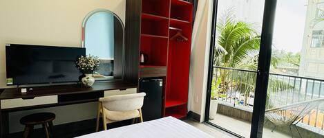 A hotel room with a bed, a desk with a TV, a red wardrobe, and a balcony with a view of palm trees.