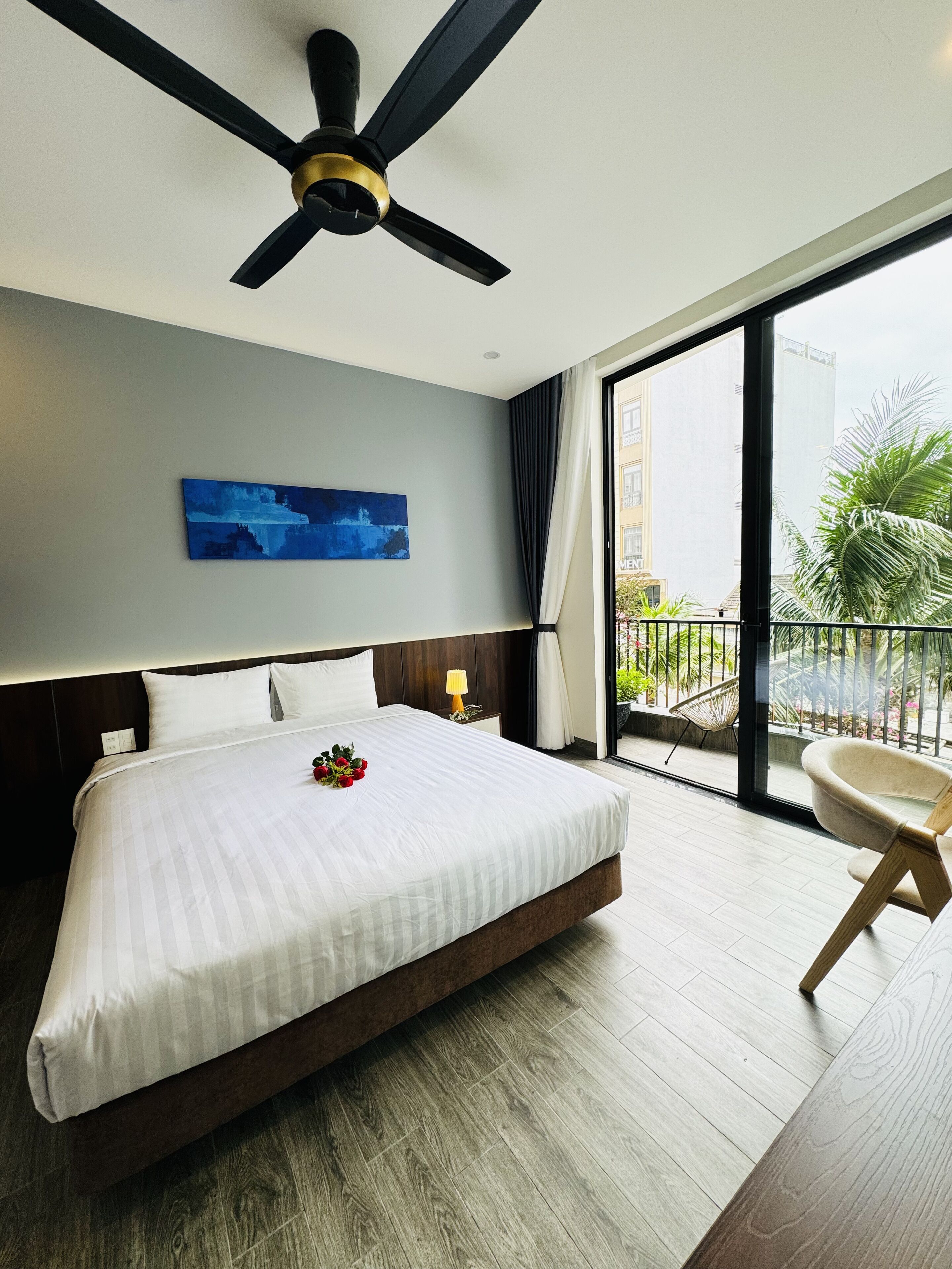 A hotel room with a large bed, a ceiling fan, a view of the city, and a balcony with a chair.