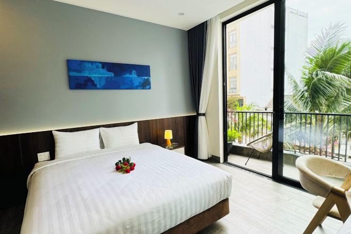 A hotel room with a large bed, a view of the outdoors, a painting on the wall, and a chair by the window.