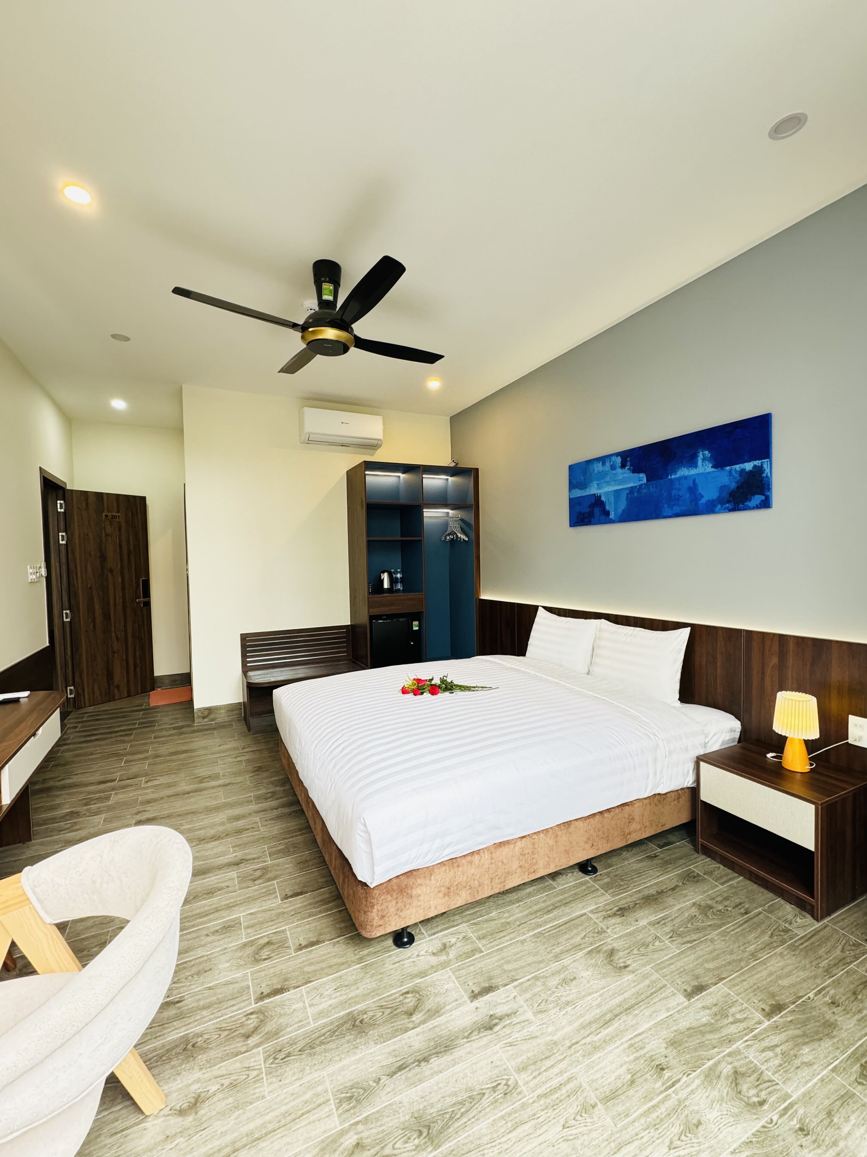 A modern hotel room with a bed, bedside table, chair, wardrobe, and ceiling fan.