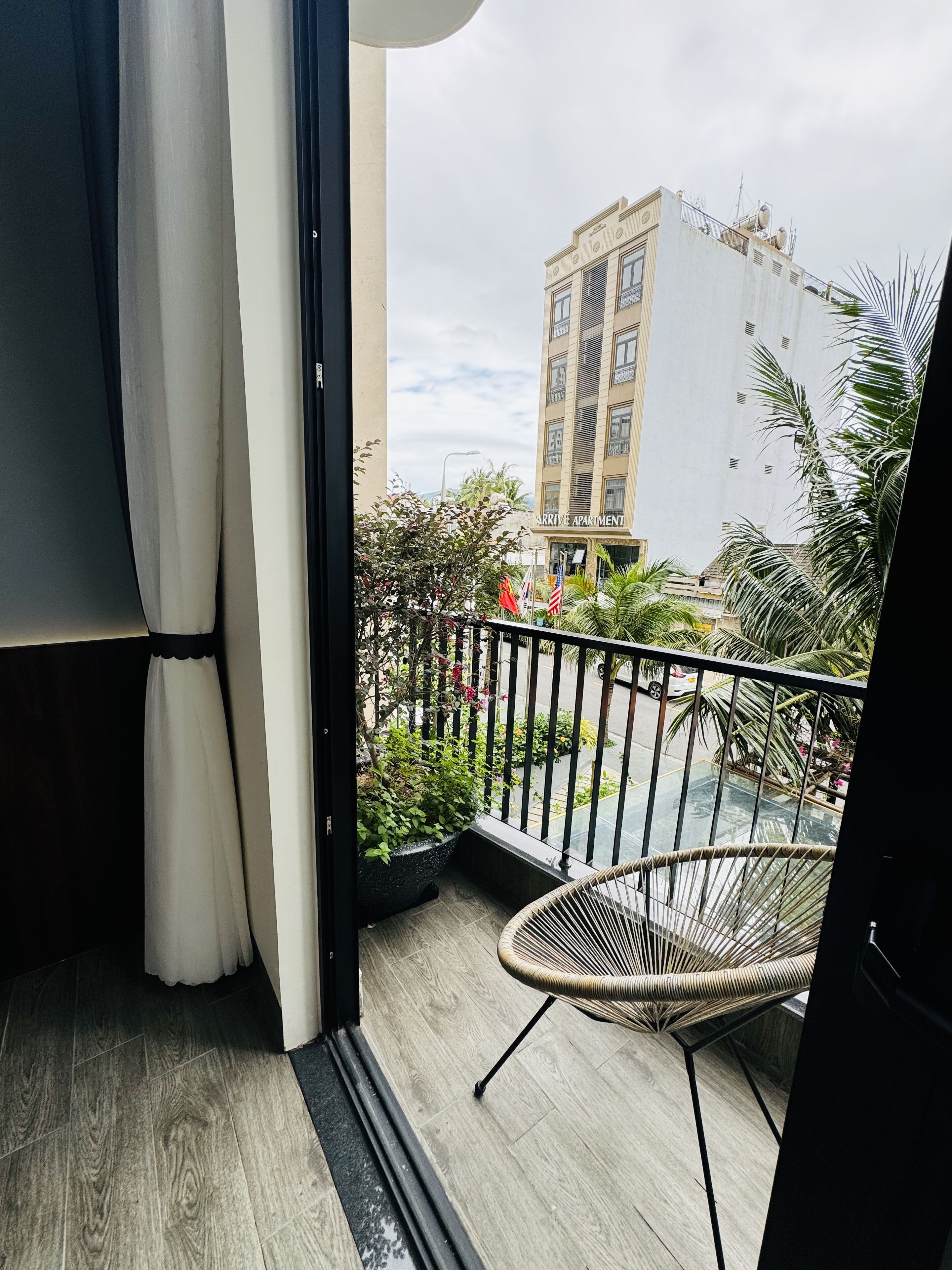 A balcony with a wicker chair, a potted plant, and a view of a building labeled "ARRIVE APARTMENT".