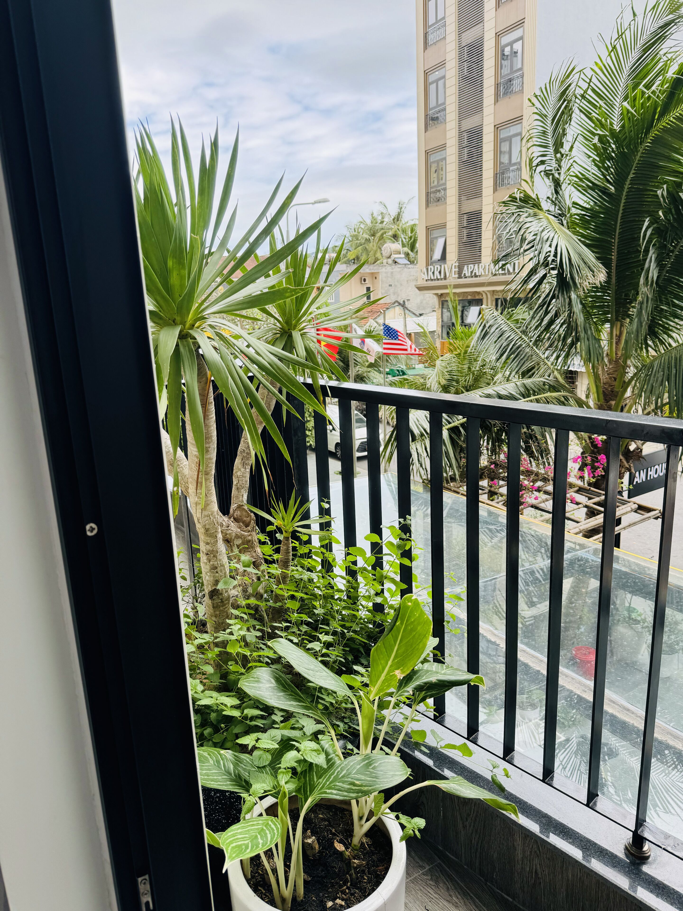 A balcony with potted plants, a view of a building labeled "ARRIVAL APARTMENT," and a street with a red car.