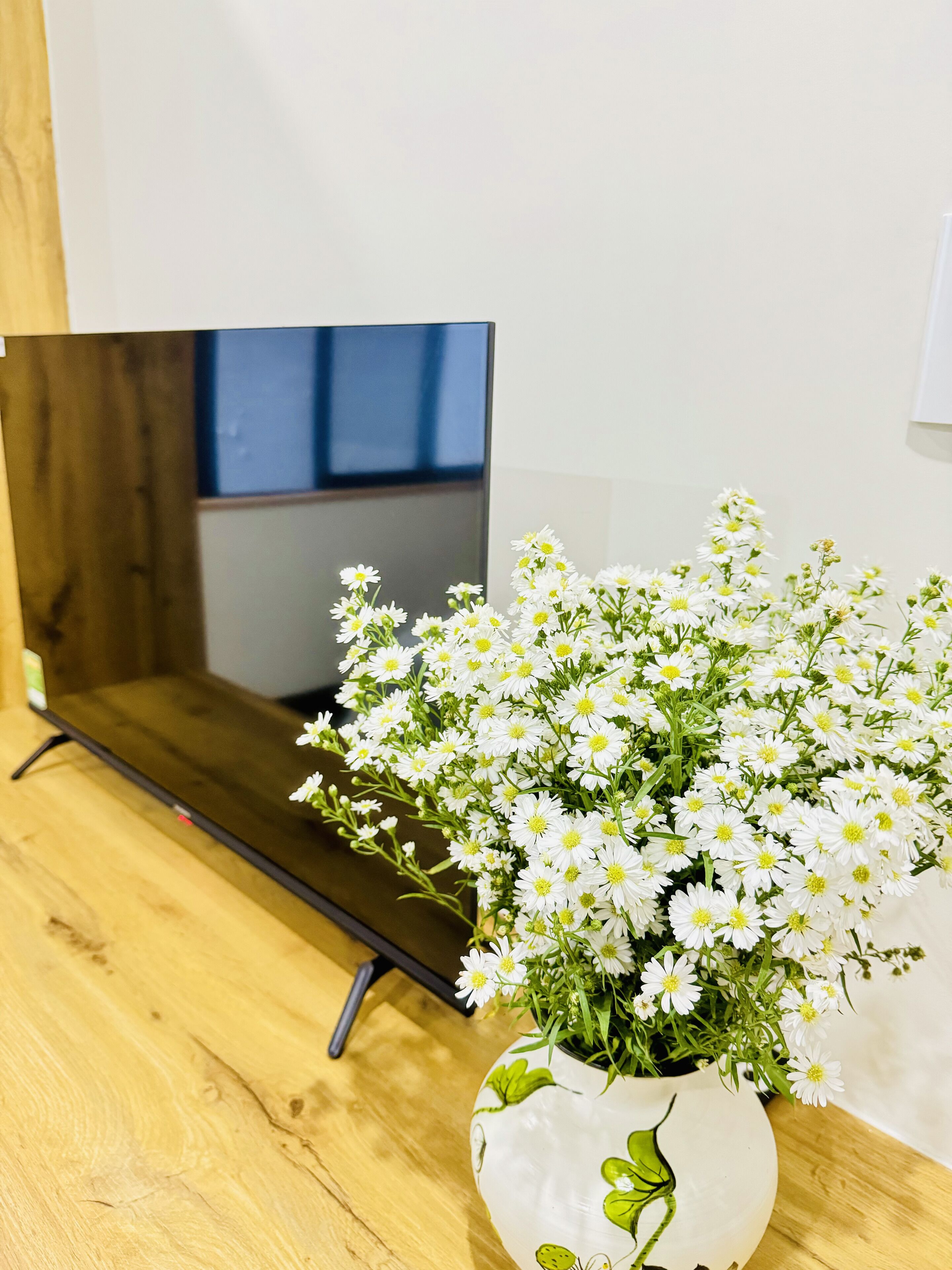 Een witte vaas met groene bladmotieven, gevuld met witte bloemen met gele kernen, staat op een houten oppervlak naast een donkere tv-meubel.