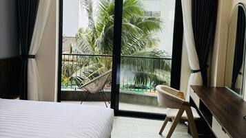 A hotel room with a bed, a desk, a chair, a large window with a view of palm trees and buildings, and a ceiling fan.