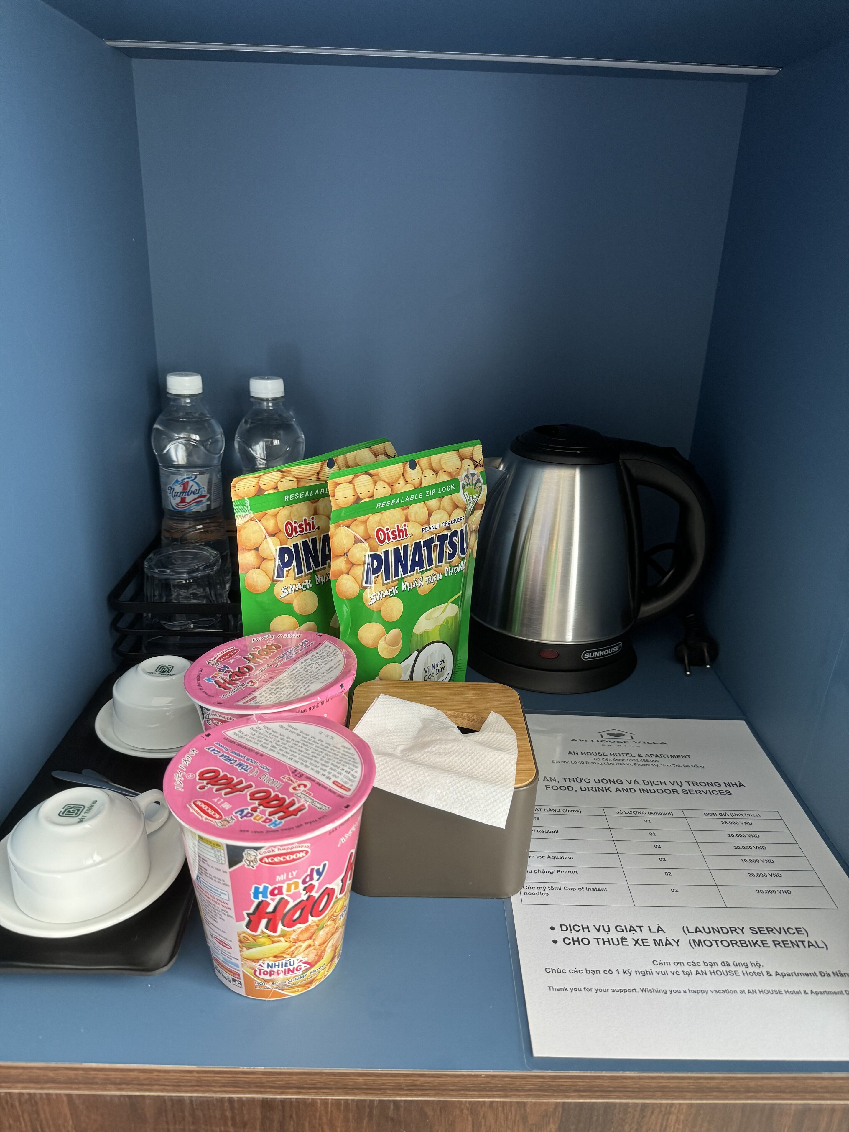 A kitchenette with a kettle, water bottles, snack packages, a coffee maker, and a menu.