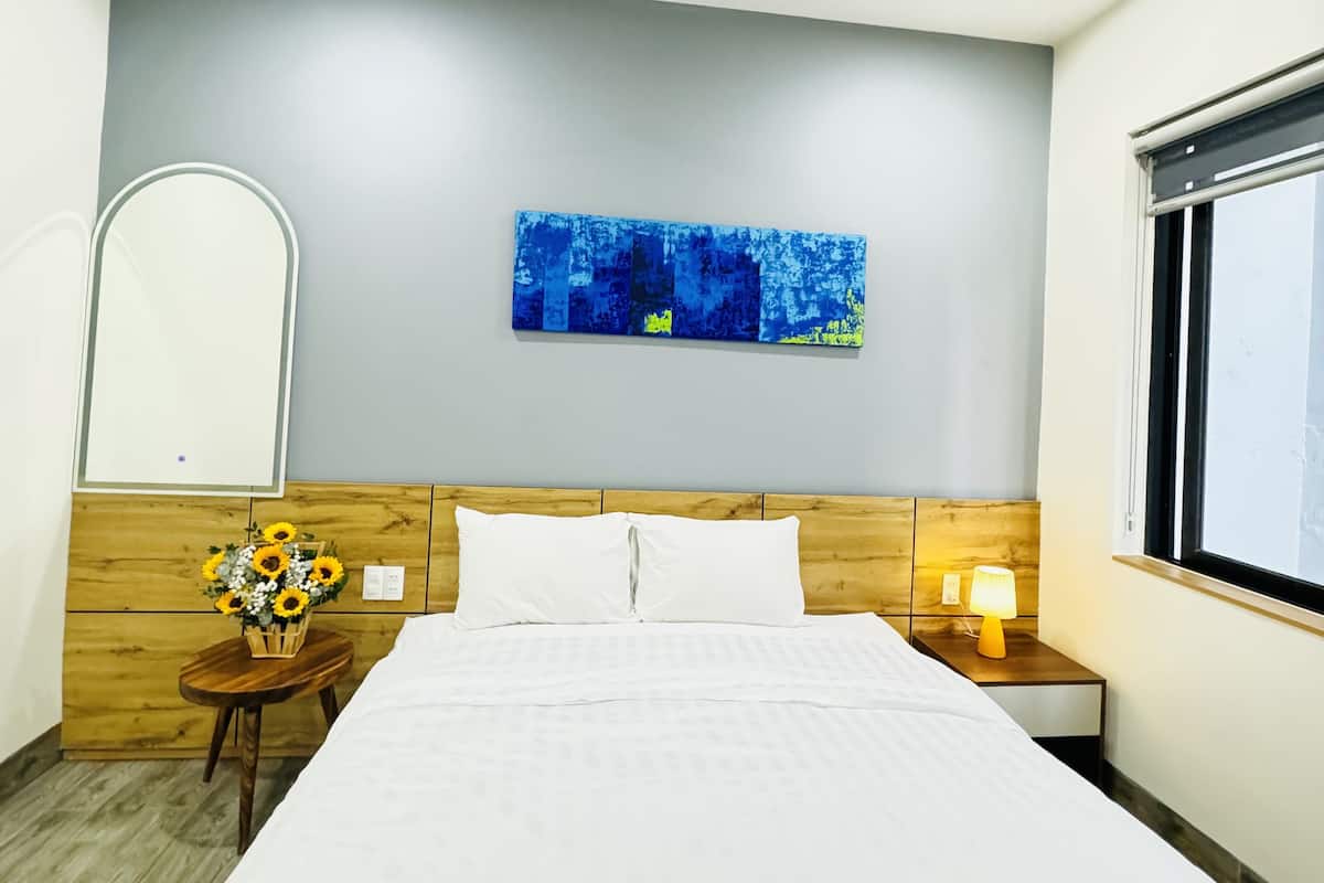 A hotel room with a bed, a ceiling fan, a wall-mounted artwork, a bedside table with a lamp, and a window.