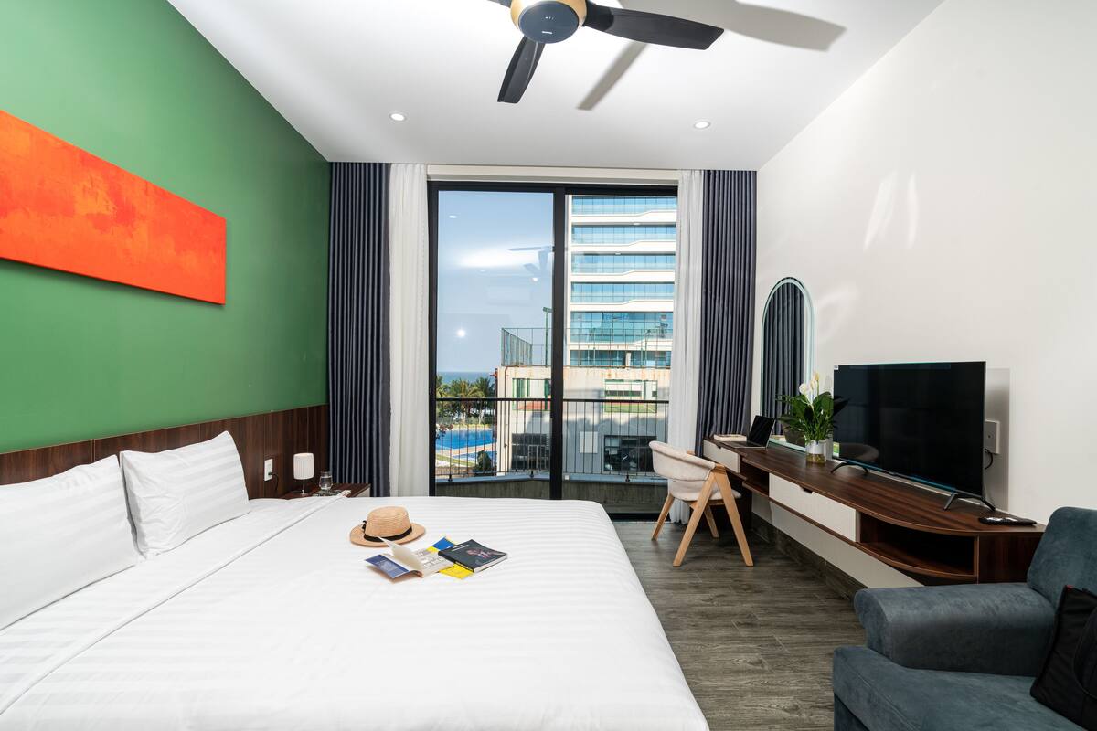 A hotel room with a bed, a desk, a TV, and a view of buildings and a pool.