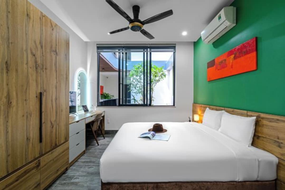 A hotel room with a bed, a desk, a wardrobe, a ceiling fan, and a window with a view of greenery.