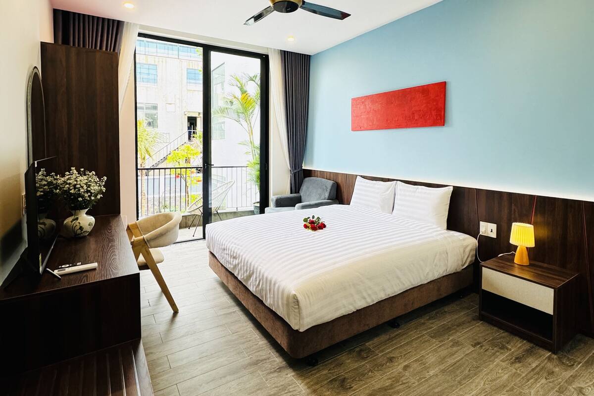 A modern hotel room with a large bed, a desk with a flat-screen TV, a ceiling fan, and a view of the outdoors.