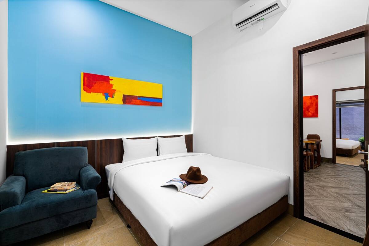 A hotel room with a large bed, a blue accent wall, a modern chair, and a view into another room.