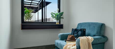 Ein Zimmer mit einem türkisblauen Sessel, einem Fenster mit Blick auf ein Gebäude und Pflanzen sowie einem Holzboden.