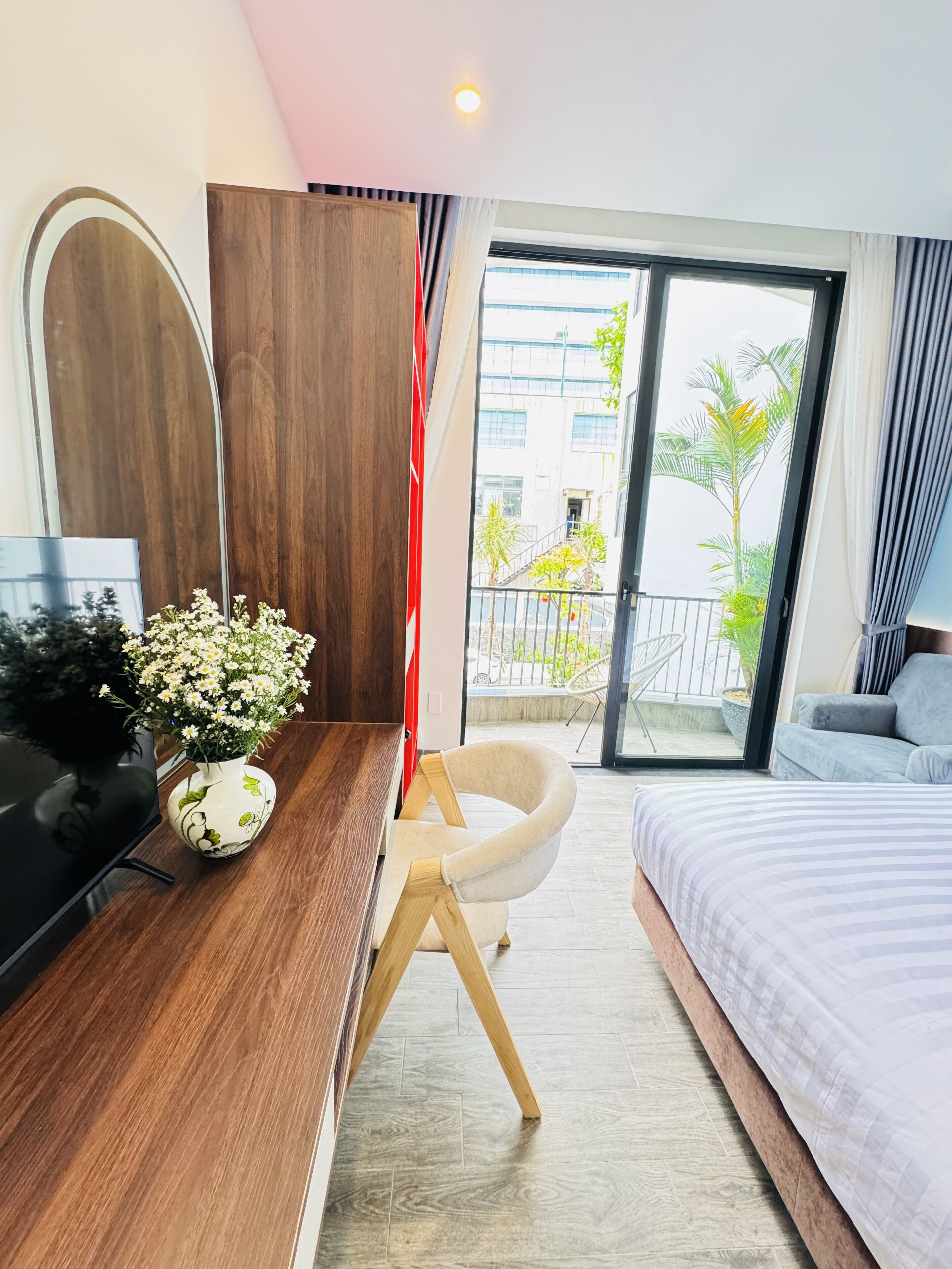 Habitación de hotel con cama, escritorio con computadora, silla, florero con flores y vista al balcón exterior.
