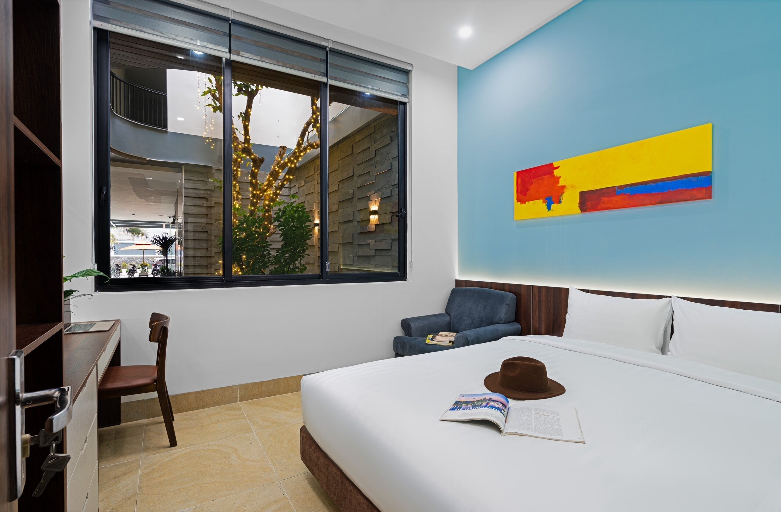 A hotel room with a bed, a desk, a chair, a window with a view of the outside, and a wall with a colorful abstract painting.