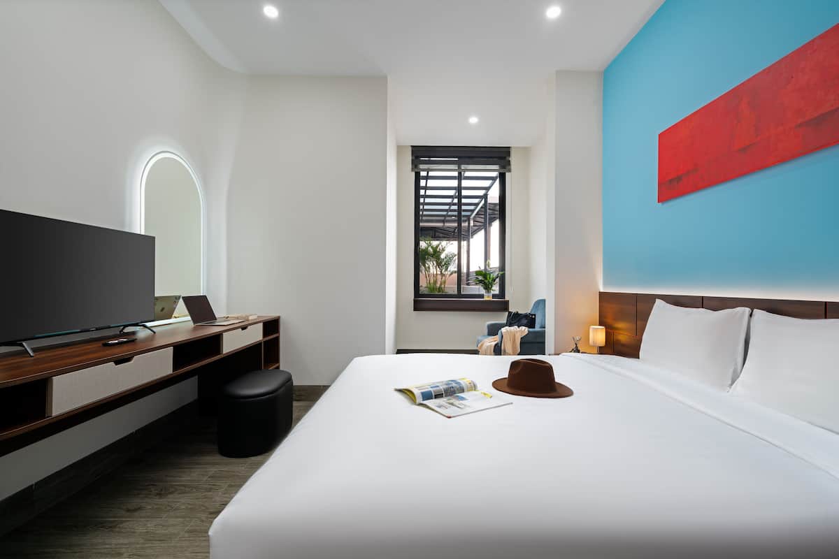 A modern hotel room with a large bed, a desk with a laptop, a television, and a window with a view of the outdoors.