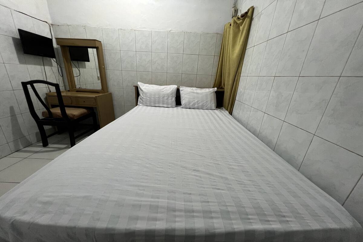A small, clean room with a bed, a wooden chair, a mirror, and a television mounted on the wall.