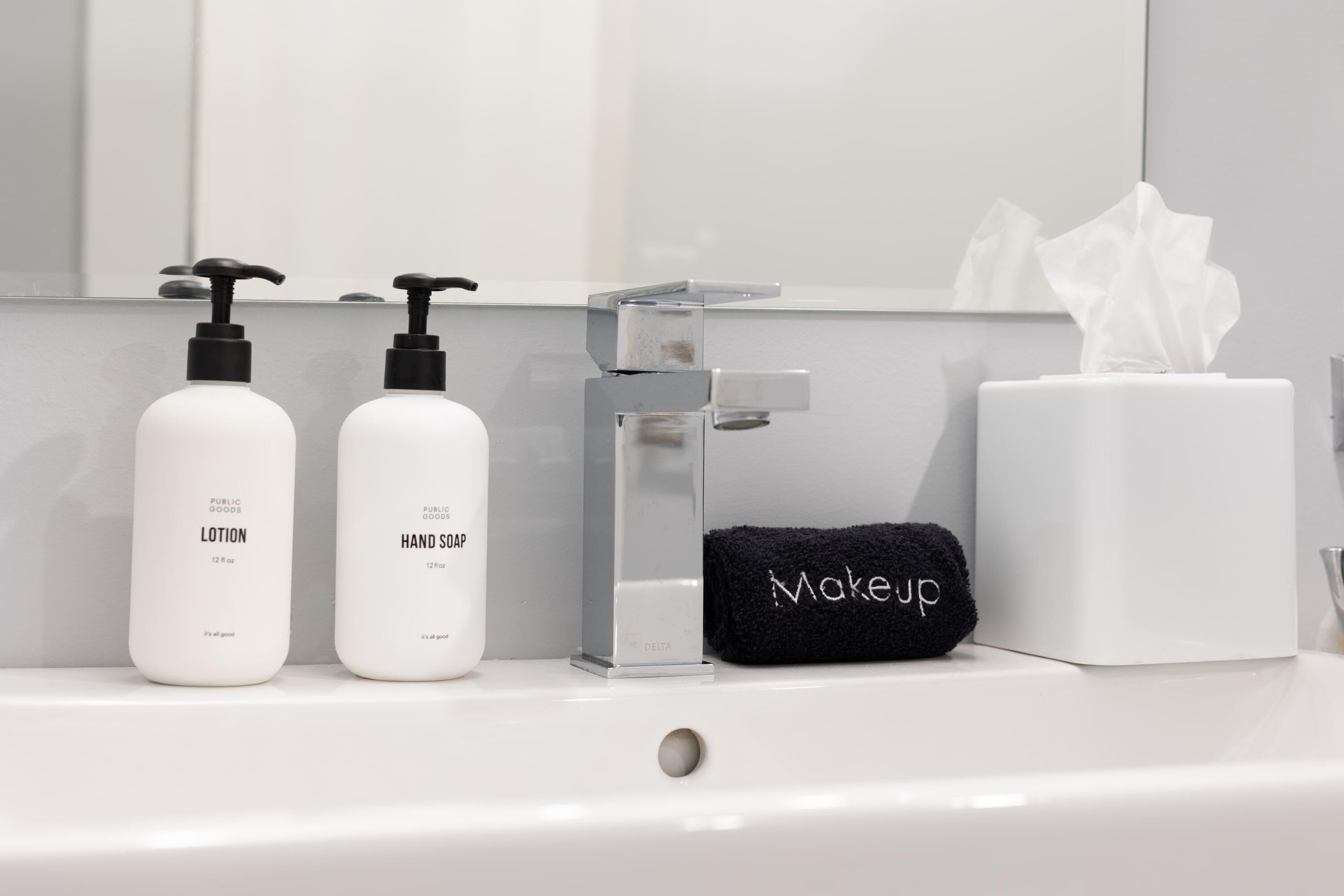 A bathroom sink with two bottles of lotion and hand soap, a clear soap dispenser, a black towel with "Makeup" written on it, and a tissue box.