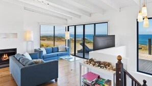 A spacious living room with a large window, a fireplace, a blue sofa, a coffee table, and a bookshelf. - Ultimate Beachfront Retreat (Sunderland Bay)