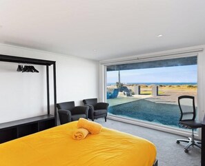 A hotel room with a bed, a desk, two chairs, and a large window with a view of the beach. - Ultimate Beachfront Retreat (Sunderland Bay)