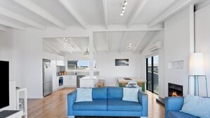 A modern living room with a blue sofa, a fireplace, and a kitchen in the background. - Ultimate Beachfront Retreat (Sunderland Bay)